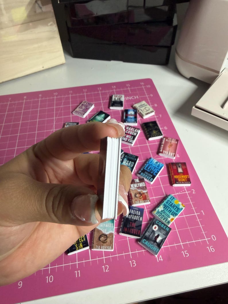 May include: A collection of miniature book replicas with various book titles visible, including "It Starts With Us" and "The Housemaid's Secret." The books are scattered on a pink cutting mat, with a stack of books held in a hand.