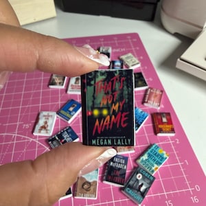 May include: Miniature book titled "That's Not My Name" by Megan Lally, held by a person. The book has a black cover with red and yellow text. Other miniature books are scattered around on a pink cutting mat.
