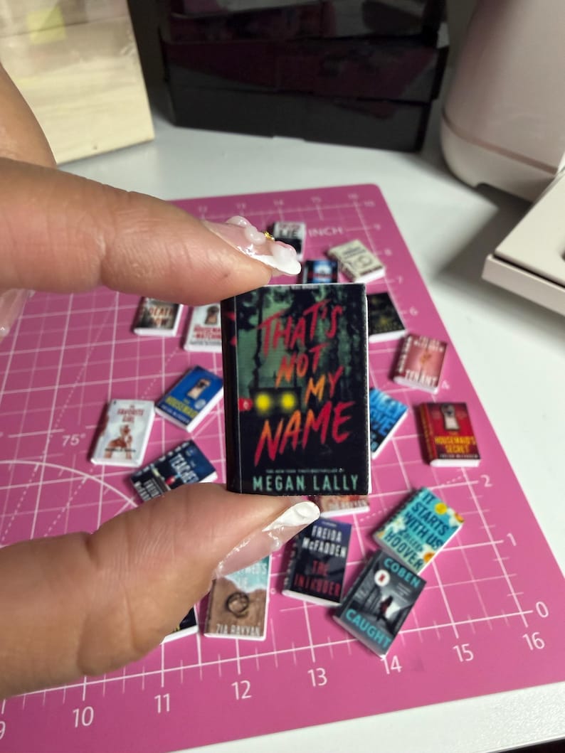May include: Miniature book titled "That's Not My Name" by Megan Lally, held by a person. The book has a black cover with red and yellow text. Other miniature books are scattered around on a pink cutting mat.