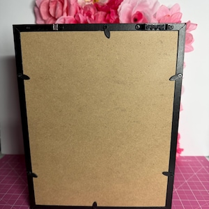 May include: Back of a black framed picture with a brown cardboard backing. The frame has metal clips to hold the backing in place. Pink and red flowers are arranged at the top of the frame, adding a decorative touch.