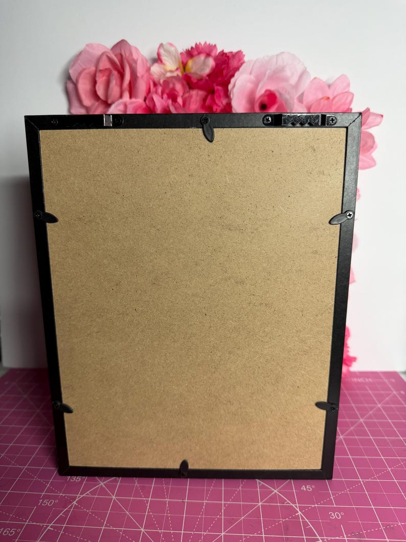 May include: Back of a black framed picture with a brown cardboard backing. The frame has metal clips to hold the backing in place. Pink and red flowers are arranged at the top of the frame, adding a decorative touch.