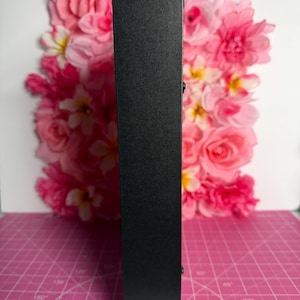 May include: A tall, black, rectangular object with a textured surface. The object is positioned vertically against a backdrop of pink and white flowers. The background is a pink cutting mat with degree markings.