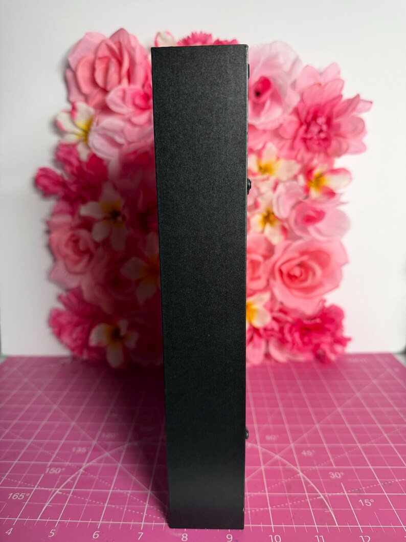 May include: A tall, black, rectangular object with a textured surface. The object is positioned vertically against a backdrop of pink and white flowers. The background is a pink cutting mat with degree markings.