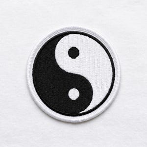 Könnte beinhalten: Ein kreisförmiger, gestickter Aufnäher mit dem Yin-und-Yang-Symbol. Das Design ist in zwei gebogene Abschnitte unterteilt, einen schwarzen und einen weißen, mit einem kleinen weißen Punkt im schwarzen Abschnitt und einem kleinen schwarzen Punkt im weißen Abschnitt. Der Aufnäher hat einen weißen, genähten Rand.