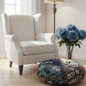 Könnte beinhalten: Ein cremefarbener Ohrensessel mit dunklen Holzbeinen. Ein runder, gemusterter Hocker mit floralen Designs steht vor dem Stuhl. Eine Vase mit blauen Blumen steht neben dem Stuhl.