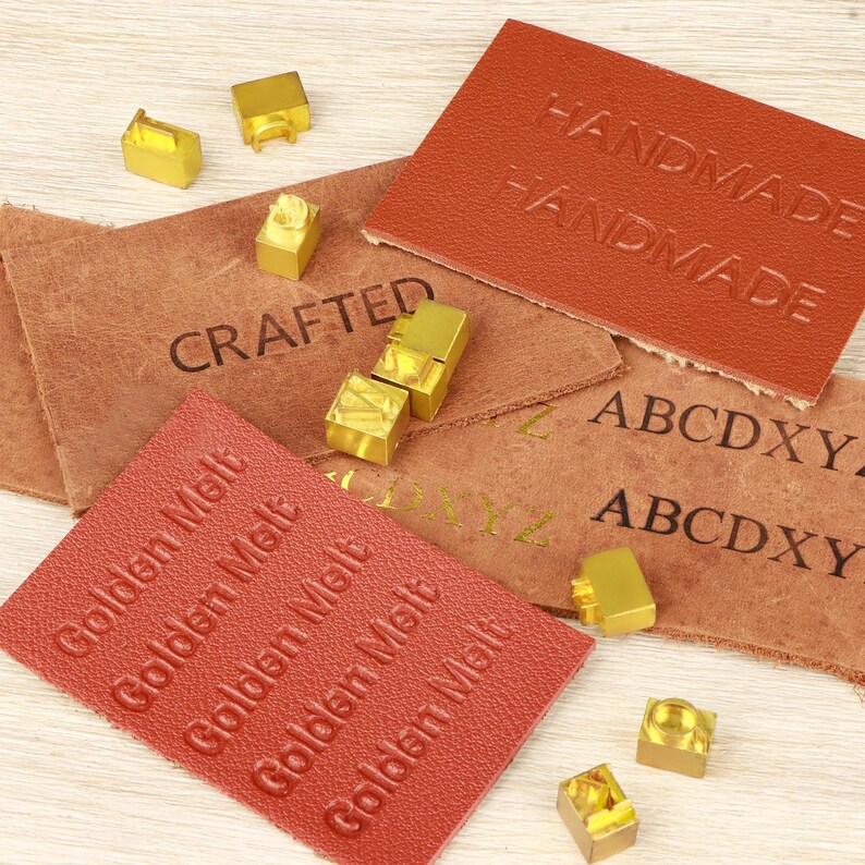 May include: An assortment of leather crafting supplies, including embossed leather tags with the words "HANDMADE", "CRAFTED", and "Golden Melt". Also, there are several small, gold-colored metal stamps and letter blocks.