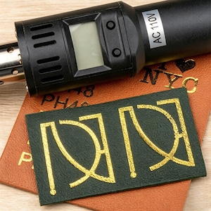 Op de afbeelding: Een zwart hitte-embossinggereedschap met een AC 110V-label en digitaal display boven een bruin leren stuk met NYC-tekst en een donkergroene rechthoekige leren patch met een goudkleurig gestempeld geometrisch logo.