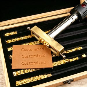 May include: A brass branding iron tool with a heating element and a metal holder for interchangeable letters, positioned over a tan leather patch embossed with the word Customized three times, all stored in a wooden case with black velvet slots.
