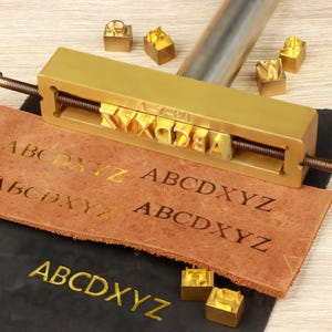 Könnte beinhalten: Ein Messing-Buchstabenstempelwerkzeug mit den Buchstaben "ABCDXYZ" auf einem Stück braunem Leder. Das Werkzeug enthält einzelne Buchstabenstempel und einen Metallgriff. Der Text "1-7-80" ist sichtbar.