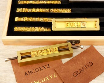 May include: A gold-colored metal branding iron tool with adjustable screws, featuring a set of interchangeable brass letter stamps arranged in a wooden storage box with black velvet lining, alongside two brown leather swatches embossed with the text ABCDXYZ and CRAFTED.
