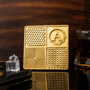 May include: A square, gold-colored coaster with four distinct textured patterns. One section features a honeycomb design, another a geometric pattern, a third a wavy pattern, and the last a circular emblem with the letter "A". Ice cubes and a glass of amber liquid are in the background.