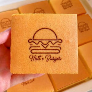 Peut inclure: Une pâtisserie carrée, orange clair, avec un logo de burger marron foncé et le texte "Matt's Burger" imprimé sur le dessus. D'autres pâtisseries avec le même logo sont visibles en arrière-plan.