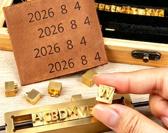 May include: A set of brass letter and number stamps for leather embossing. The kit includes a metal holder with a threaded rod, individual brass character blocks, and a wooden storage box. A square piece of brown leather shows the stamped text 2026 8 4.