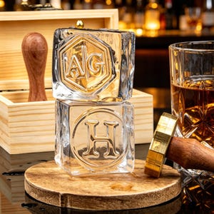 May include: Two clear, square ice cubes stacked vertically, each featuring a debossed letter design. The top cube displays the letters AG inside a hexagon, while the bottom cube features the letter H inside a circle. A wooden handle tool sits nearby.