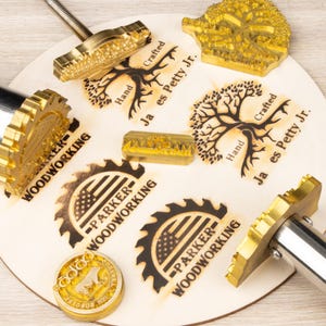 Puede incluir: Un conjunto de sellos y hierros de marcar de latón para carpintería. Los sellos incluyen diseños de árboles y hojas de sierra, junto con el texto "PARKER WOODWORKING" y "Hand Crafted Ja es Petty Jr.". Dispuestos sobre una superficie de madera clara.