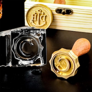 May include: A brass wax seal stamp featuring a letter C, with a wooden handle and a scalloped edge. Beside it, a clear glass block shows a stamped impression of the letter C. A wooden box with a metal latch and a round brass seal are visible.