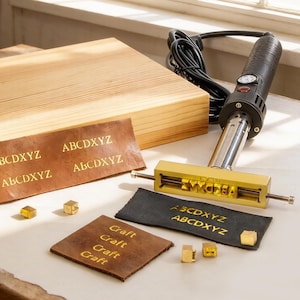 May include: A leathercrafting set featuring a branding iron, wooden block, and various leather samples. The samples display stamped text, including "ABCDXYZ" and "Craft". Gold-colored metal letter cubes are also present, suggesting a customizable design process.