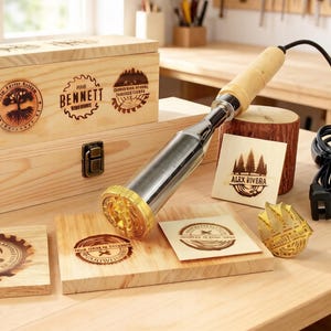 Puede incluir: Un hierro de marcar madera con mango de madera clara y elemento calefactor metálico con cabezal de sello circular dorado. La herramienta se muestra junto a una caja de madera, varias baldosas cuadradas y un tronco, todos marcados con diseños circulares quemados que incluyen el texto BENNETT y ALAX RIVERA.