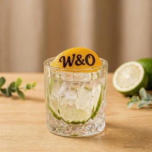 May include: A patterned glass filled with a refreshing drink, ice cubes, and lime slices. A yellow orange slice with the letters "W&O" sits on top. The drink is on a wooden surface with a blurred background.