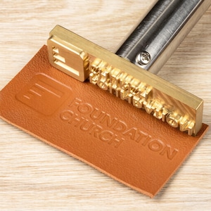 Puede incluir: Un sello de latón con las palabras "FOUNDATION CHURCH" grabadas en relieve sobre un trozo de cuero marrón. El sello tiene un mango plateado y está presionado sobre el cuero, creando una impresión en relieve. El cuero tiene forma rectangular.