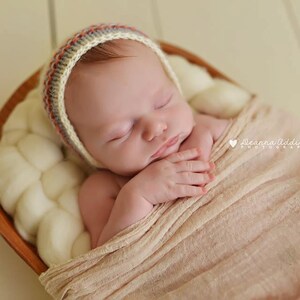 May include: A newborn baby wearing a knitted bonnet with red, gray, and orange stripes, wrapped in a peach-colored blanket, and nestled in a wooden bowl filled with soft, white material. The baby is sleeping peacefully.