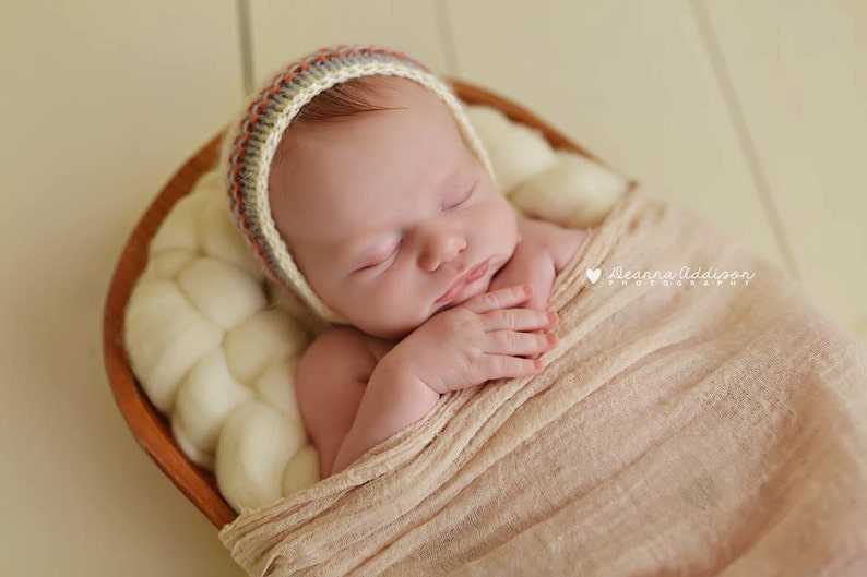 May include: A newborn baby wearing a knitted bonnet with red, gray, and orange stripes, wrapped in a peach-colored blanket, and nestled in a wooden bowl filled with soft, white material. The baby is sleeping peacefully.