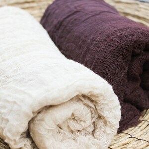 May include: Two rolled-up linen scarves. One is a creamy off-white, the other a deep burgundy. The fabric has a crinkled texture. The scarves are displayed on a woven, circular mat, suggesting a natural, rustic aesthetic.