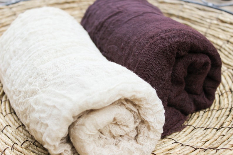 May include: Two rolled-up linen scarves. One is a creamy off-white, the other a deep burgundy. The fabric has a crinkled texture. The scarves are displayed on a woven, circular mat, suggesting a natural, rustic aesthetic.