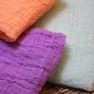 May include: Close-up of three pieces of fabric in shades of orange, purple, and light green. The fabric has a crinkled, textured appearance, suggesting a gauze or cheesecloth material. The colors are soft and muted.