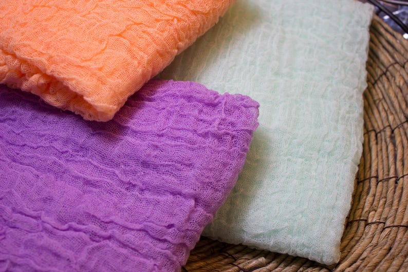 May include: Close-up of three pieces of fabric in shades of orange, purple, and light green. The fabric has a crinkled, textured appearance, suggesting a gauze or cheesecloth material. The colors are soft and muted.