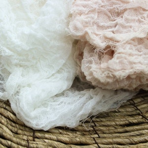 May include: Close-up of two pieces of textured fabric, one white and one light pink, draped over a woven basket. The fabrics have a crinkled, gauze-like appearance, suggesting a soft, delicate texture. The basket is made of natural, brown fibers.
