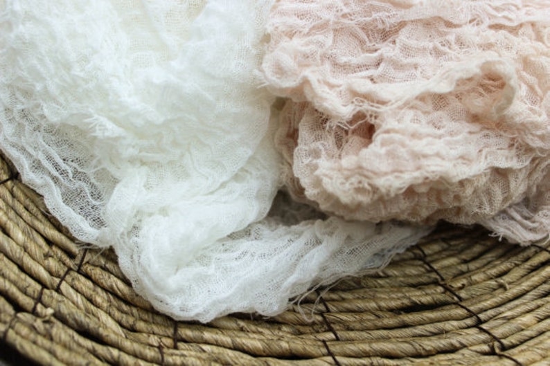 May include: Close-up of two pieces of textured fabric, one white and one light pink, draped over a woven basket. The fabrics have a crinkled, gauze-like appearance, suggesting a soft, delicate texture. The basket is made of natural, brown fibers.