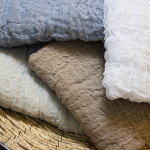 May include: Close-up of several folded, crinkled fabric cloths in shades of blue, white, and tan. The cloths are stacked inside a woven basket. The fabric has a textured, gauzy appearance.