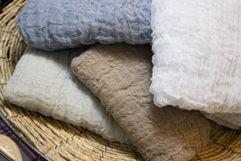May include: Close-up of several folded, crinkled fabric cloths in shades of blue, white, and tan. The cloths are stacked inside a woven basket. The fabric has a textured, gauzy appearance.