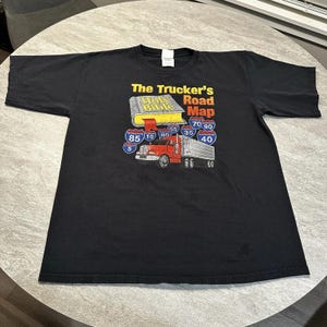 May include: Black t-shirt with a graphic of a red lorry with a trailer, a yellow book labelled "Holy Bible", and road signs. The text reads "The Trucker's Road Map."