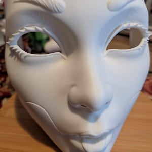 May include: A white, blank mask with detailed facial features. The mask has sculpted eyebrows, large eye openings with spiky lashes, a nose, and a smiling mouth. The mask is made of a smooth, matte material.
