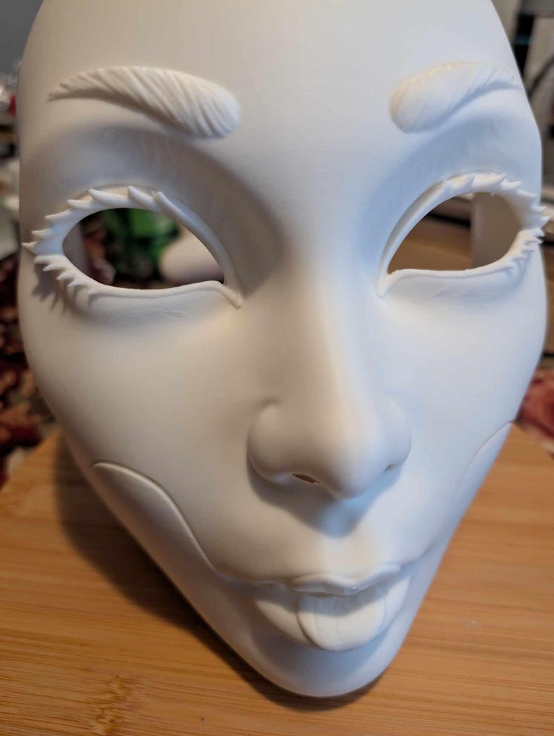 May include: A white, blank mask with detailed facial features. The mask has sculpted eyebrows, large eye openings with spiky lashes, a nose, and a smiling mouth. The mask is made of a smooth, matte material.