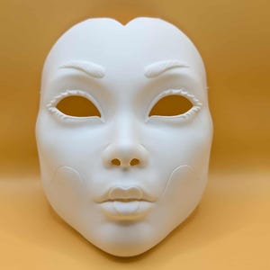 May include: A blank, white mask with a human face design. The mask features eye holes, a nose, and a mouth. The eyebrows and eyelashes are also detailed. The mask is likely for costume or decorative purposes.