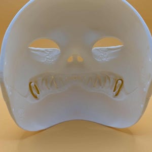 May include: A white, molded plastic mask with eye and nose openings. The mask has a wide, tooth-like design along the bottom edge and two small, curved metal pieces on either side of the mouth.