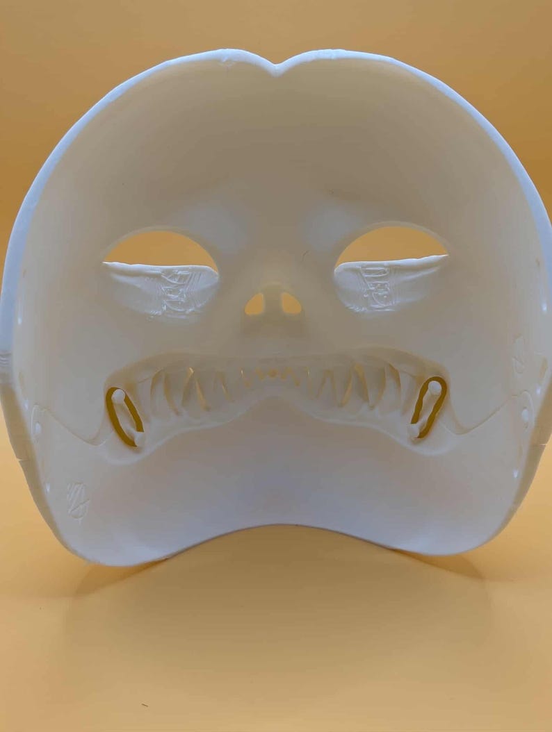May include: A white, molded plastic mask with eye and nose openings. The mask has a wide, tooth-like design along the bottom edge and two small, curved metal pieces on either side of the mouth.