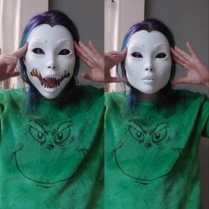 May include: Two white masks, one with a detailed open-mouth design revealing teeth, and the other with a closed mouth. The masks are worn over the face. The person is wearing a green sweatshirt with a Grinch graphic.