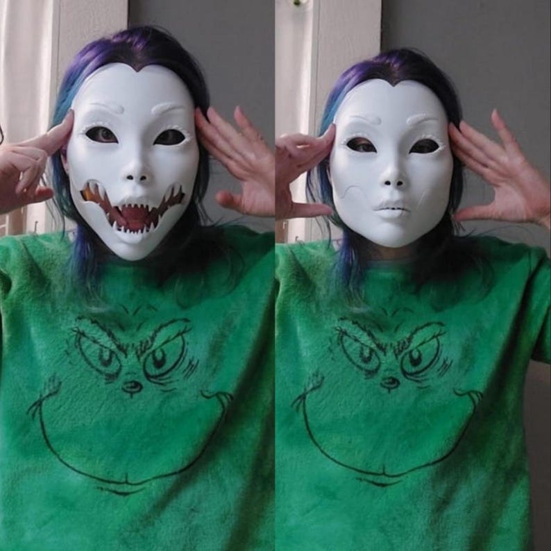 May include: Two white masks, one with a detailed open-mouth design revealing teeth, and the other with a closed mouth. The masks are worn over the face. The person is wearing a green sweatshirt with a Grinch graphic.