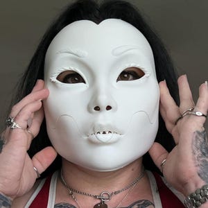 May include: A white mask with a serene expression, featuring delicate facial contours and eye cutouts. The mask is held by hands adorned with rings and tattoos. The person's eyes are visible through the eye cutouts. The mask is a costume or decorative item.
