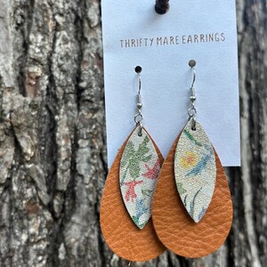 Peut inclure: Boucles d'oreilles en forme de goutte sur un pr&eacute;sentoir blanc marqu&eacute; THRIFTY MARE EARRINGS. Chaque boucle pr&eacute;sente une grande base en cuir beige textur&eacute; superpos&eacute;e d'un insert en tissu blanc plus petit orn&eacute; d'un motif floral color&eacute;.