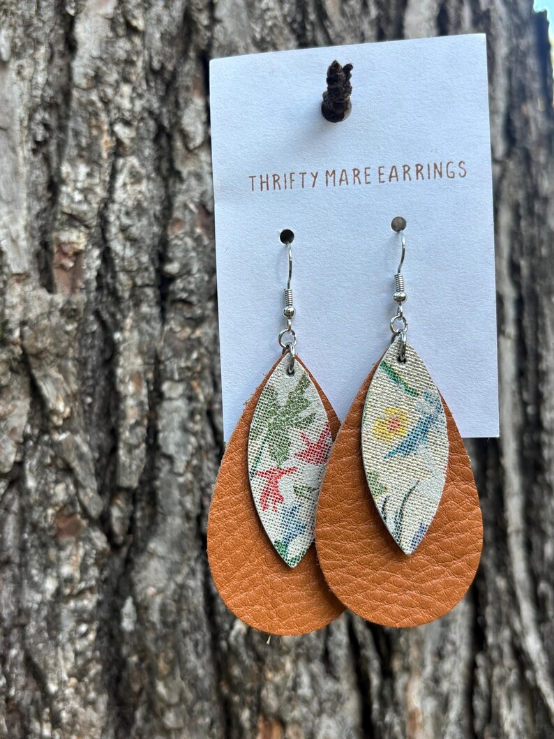 Peut inclure: Boucles d'oreilles en forme de goutte sur un pr&eacute;sentoir blanc marqu&eacute; THRIFTY MARE EARRINGS. Chaque boucle pr&eacute;sente une grande base en cuir beige textur&eacute; superpos&eacute;e d'un insert en tissu blanc plus petit orn&eacute; d'un motif floral color&eacute;.