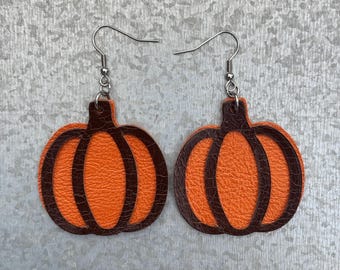 Peut inclure: Une paire de boucles d'oreilles en forme de citrouille avec une matière texturée orange semblable à du cuir superposée sous un cadre découpé marron foncé. Elles pendent à des crochets en métal argenté.