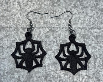 Peut inclure: Une paire de boucles d'oreilles pendantes noires présentant un motif découpé d'une araignée au centre d'une toile. Les boucles d'oreilles sont suspendues à des crochets en métal argenté, avec une finition mate et texturée.