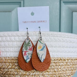 Peut inclure: Une paire de boucles d'oreilles en forme de goutte avec un design superpos&eacute;, comprenant une base en cuir textur&eacute; marron uni et une couche sup&eacute;rieure plus petite et claire orn&eacute;e d'un motif floral color&eacute;. Elles sont fix&eacute;es sur une carte blanche marqu&eacute;e THRIFTY MARE EARRINGS.