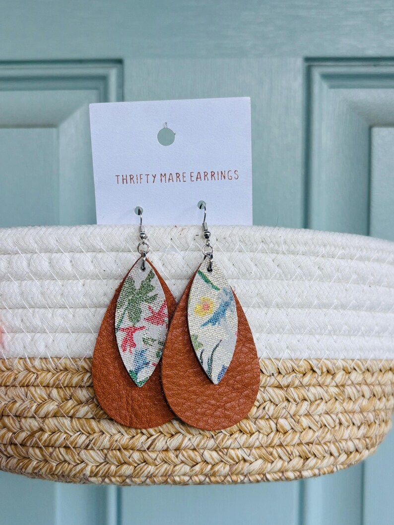 Peut inclure: Une paire de boucles d'oreilles en forme de goutte avec un design superpos&eacute;, comprenant une base en cuir textur&eacute; marron uni et une couche sup&eacute;rieure plus petite et claire orn&eacute;e d'un motif floral color&eacute;. Elles sont fix&eacute;es sur une carte blanche marqu&eacute;e THRIFTY MARE EARRINGS.
