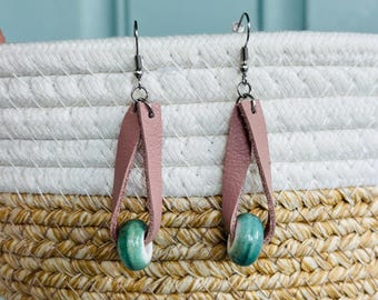 Peut inclure: Une paire de boucles d'oreilles pendantes composées d'une lanière de cuir rose poudré en forme de boucle. Chaque boucle en cuir retient une perle ronde en céramique émaillée avec un dégradé de blanc et de vert sarcelle, fixée à des crochets argentés.